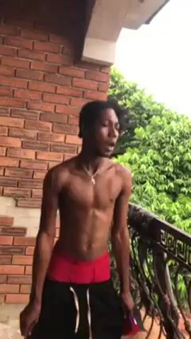 A shirtless, slim, athletic man with dark hair, red briefs, and black baggy pants dances outside on a patio of some kind. He holds a red hat in his left hand. The patio is made of a light tan brick. There is a window to the left of the man. There are green leafy trees in the background.