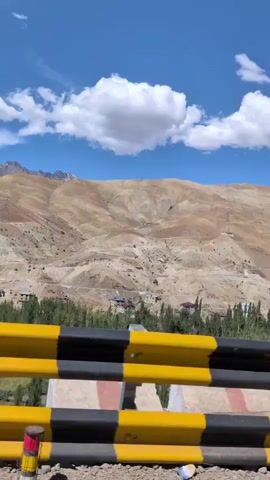 The video captures a scenic view of a mountainous region under a bright blue sky with scattered white clouds. A yellow and black striped barrier is prominently in the foreground, suggesting the perspective is from a vehicle moving along a road. Behind the barrier, a dry, arid mountain landscape unfolds, with slopes marked by roads and some scattered structures. A few trees are visible in the mid-ground, indicating some sparse vegetation in the valley. A utility pole with wires stands on the left side, further establishing the roadside setting. The camera is in motion, creating a panning effect that showcases the vastness and rugged beauty of the terrain.