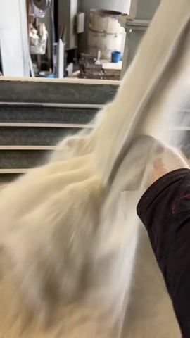 A close-up video shows a stream of fine, white powder or sand cascading over a person's outstretched hand. The hand, adorned with a dark, long-sleeved shirt, is positioned to catch the flowing material, which appears very smooth and fluid-like. In the blurred background, an industrial setting is visible, featuring various metal structures, possibly machinery components, and what looks like a large cylindrical tank or container, along with a small blue bucket. The lighting suggests an indoor environment. No text is visible.