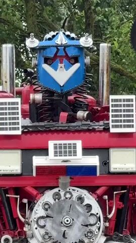 This media file features a life-size sculpture of Optimus Prime, a character from the Transformers franchise, created from recycled materials. The sculpture is predominantly red and blue, with various metal parts forming its body, including gears, pipes, and what appear to be car parts. Two solar panels are visible on the upper chest area, and two more are mounted on the shoulders. The sculpture's head is blue with a white and red faceplate.

The sculpture stands outdoors against a backdrop of lush green trees, suggesting a natural or park-like setting. The lighting indicates it's daytime, likely overcast or shaded by the trees, as there are no strong shadows. To the left of the sculpture, a vintage red and blue motorcycle is parked, also appearing to be made of recycled parts. The ground is a rough, unpaved surface. The overall impression is that of an outdoor art installation or a themed display. There is no visible text on the sculpture itself, though two small rectangular panels on the lower chest read "60W Solar Light." The sculpture is the primary subject, with the motorcycle serving as a secondary element in the scene.