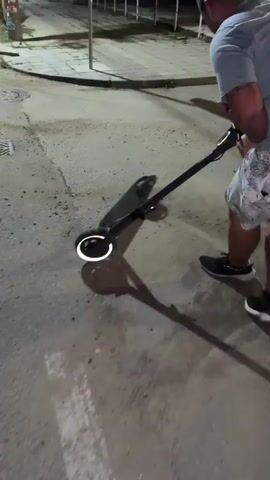 A person's legs are visible from the waist down, wearing patterned shorts and black athletic shoes. They are walking on a paved surface, possibly a street or sidewalk, at night. The person is holding the handlebars of a dark-colored electric scooter. The scooter's front wheel has a glowing white ring around it. The scooter is tilted, suggesting it's either being pushed or is about to be ridden. The ground is a mix of asphalt and gravel, with some shadows cast on it. A metal manhole cover is visible on the left side of the frame. The overall lighting suggests it's dark, with artificial light sources present.