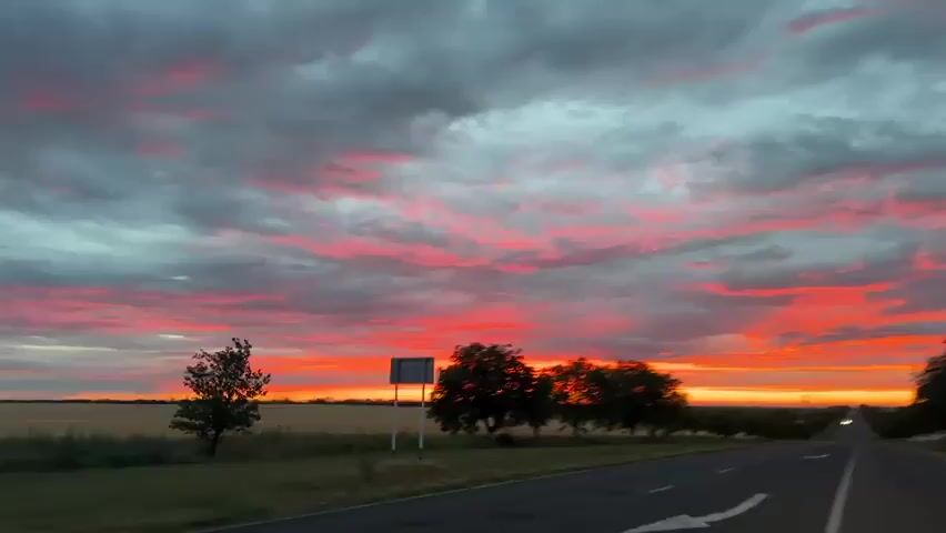 A car's dashcam captures a vivid sunset over a rural landscape. The sky is a dramatic canvas of clouds painted in shades of fiery red, orange, and gray, with the sun low on the horizon. A two-lane asphalt road stretches into the distance, bordered by fields of dry grass and a line of trees on the left side. The trees appear blurred due to the vehicle's motion. A road sign is visible on the left side of the road at the beginning of the footage. The perspective is from inside the moving vehicle, looking forward. The overall impression is one of a journey through a beautiful, albeit slightly overcast, twilight.