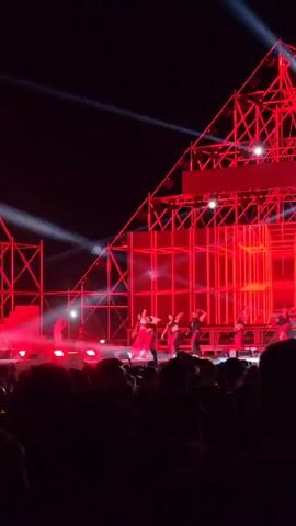 The video shows a stage performance at an outdoor concert. A group of performers, including a woman in a red outfit, are on stage, illuminated by red and white stage lights. The stage backdrop features a white screen with red lines arranged in a grid pattern, with scaffolding on either side. Below the stage, a crowd is visible, with many individuals raising their hands and recording the performance on their phones. The night sky is visible above the stage.