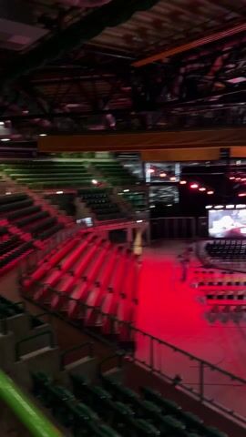 The interior of a large indoor venue is shown in this video. There is a stage set up in the center of the space with bright lights and a large screen in the background. A scattering of people are on and around the stage. Rows of empty chairs face the stage. Bleacher-style seating lines the sides of the venue. A raised walkway with a green railing is in the foreground. The venue's ceiling is high and exposed. The lighting in the venue is dim, with the stage lights providing the most illumination.