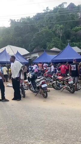 The video captures an outdoor scene on a paved road with several parked motorcycles, predominantly red and silver, on both sides. Multiple people are gathered under several blue tents and in front of buildings, including a pink structure, lining the road. A dark SUV is parked near the tents. A woman in a white jumpsuit and red sandals rides a red scooter from right to left across the frame. Subsequently, a man in a blue cap, driving a silver motorcycle, carries a woman in a yellow patterned dress as a passenger, also moving from right to left. The background features hills covered with dense foliage under an overcast sky. The overall impression is a bustling roadside gathering or event.