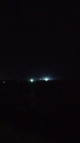 The video shows a dark, nighttime scene of a body of water, presumably the sea or a large lake. The horizon is visible, with scattered lights in the distance, possibly from buildings or ships. The water reflects some of the light, creating a shimmering effect. The overall scene is dark and still, with minimal visible detail. There are no people or distinct objects visible beyond the lights in the distance.