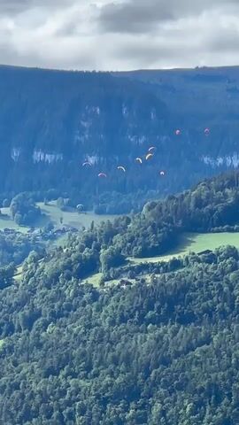 The video shows an aerial view of a mountainous landscape with dense green forests covering the slopes. Several paragliders, distinguished by their colorful canopies, are visible in the sky, spread out across the frame against a backdrop of hazy mountains and a cloudy sky. The paragliders appear to be moving and gliding over the terrain. Small buildings and fields are intermittently visible amidst the trees on the lower slopes, suggesting a rural or mountainous settlement. The overall impression is one of a recreational activity taking place in a scenic natural environment.