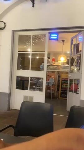 The video opens with a view of the entrance to a cafe or restaurant. The door is slightly ajar, revealing tables and chairs inside, along with some illuminated signs and decor. The exterior area in the foreground appears to be a patio or covered walkway, with dark chairs and a table visible.

As the video progresses, the camera pans slightly to the left, focusing on a man with dark hair, who is looking down and holding a cigarette. He is seated at a table.

The camera then moves to the right, showing a young woman with dark hair, wearing a red tank top, sitting at a table. She is looking towards the camera and smiling. A white and beige purse with a chain strap rests on the table in front of her.

Further to the right, a woman with blonde hair, wearing a red shirt with a "España" logo, is partially visible. She is also seated at a table. Another person, possibly male, is seen sitting in the background, wearing a dark shirt and a baseball cap, looking away from the camera. The overall environment suggests an outdoor seating area of a cafe or restaurant, possibly in the evening or late afternoon, given the lighting.