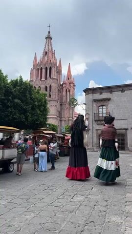 The video shows a stone-paved plaza with people walking around, food carts, and two large standing figures in traditional Mexican clothing. In the background, there is a tall, ornate church and a stone building. The sky is cloudy, and there are some trees visible on the left side of the frame.