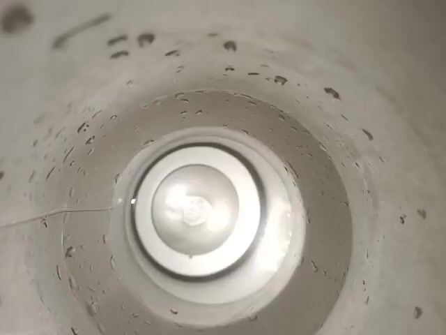 The video shows a close-up, interior view of a white cylindrical container, possibly a mug or a thermos. The camera appears to be looking down into the container. The inner walls are slightly textured and have several small water droplets clinging to them. Towards the bottom, a circular, raised surface is visible, with concentric rings. A bright light source is reflected in the center of this surface, creating a luminous glow. The overall impression is of looking into an empty or nearly empty container that has recently held liquid.