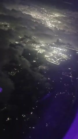 The video shows a night aerial view of a city at night from an airplane window. The city lights are visible through clouds, creating a glowing effect. A large brightly lit area, possibly an airport or industrial complex, is prominent. The purple tint at the bottom right suggests the interior of the airplane cabin is visible. The camera is positioned to look down and slightly ahead.