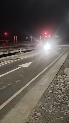 The video shows a night-time view of a toll booth. A car with its headlights on approaches the toll booth, its bright lights illuminating the area. Red lights are visible on the toll booth signals, indicating that traffic should stop. The road is marked with white lines, and there is a sidewalk with scattered rocks to the right. The background is dark, suggesting it is nighttime. The camera appears to be mounted on a moving vehicle, as the scene is slightly blurred and shifts as the vehicle moves.