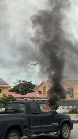 A group of individuals are gathered around a burning tire on a road. The tire is engulfed in flames, producing a large plume of dark smoke that rises into the sky. The people are standing at a distance from the fire, some appear to be observing it, while others are holding what looks like sticks. In the background, there are houses and a street lamp. The scene suggests a protest or demonstration, likely involving acts of defiance.