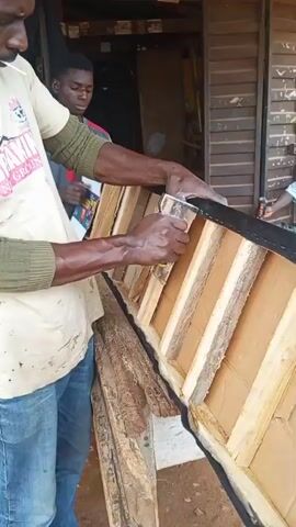 This media file captures a segment of a woodworking or upholstery project in Barikin Sabon Gidan Kanat, Nigeria. Two men are the central subjects, engaged in constructing or repairing a piece of furniture, possibly a headboard or a similar padded item.

In the foreground, a man wearing a light-colored t-shirt with graphics and blue jeans is actively working. He is holding a staple gun in his right hand, appearing to attach a black fabric or material to the wooden frame of the furniture. His left hand is steadying the material. He has a focused expression and is wearing a green long-sleeved undershirt.

Behind him, another man, younger, is also observing the process or assisting in some capacity. He is wearing a grey and red t-shirt.

The furniture piece itself is constructed with a visible wooden frame, featuring vertical slats. It is partially covered with the black material along its edge, secured by staples. A roll of black material is also visible on the ground.

The setting appears to be an outdoor workshop or a covered open-air space, suggested by the natural light and the rustic wooden structure in the background. The ground is earthy.

Notable details include the focus on the manual labor involved in attaching the fabric, indicating a hands-on construction process. The time of day appears to be daytime, with ample natural light.

Visible text on the foreground subject's t-shirt includes "SHILOH" and "BREAKING NEW GROUNDS," suggesting a possible brand or slogan associated with the garment.