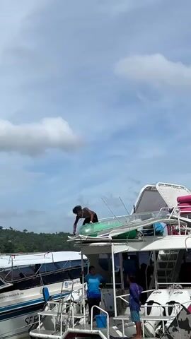 A man with dark curly hair and a necklace is leaning over the edge of a boat, holding onto a clear tube with green and white accents. He is wearing dark shorts and appears to be in motion, possibly preparing to slide down the tube. Other people are visible on the deck of the boat below him, and another boat is docked beside them. The sky is blue with white clouds, and a green, tree-covered hill is in the background.