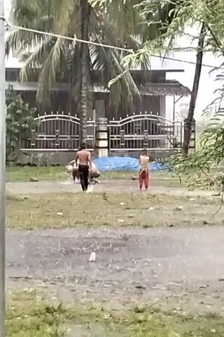 The video shows three people playing in a heavy rain on a grassy area in front of a house. Two of them appear to be shirtless. One person, wearing dark pants, is walking while the other two are crouching on the ground.  A blue tarp is laid out on the grass. The house is visible behind a gate and a large palm tree. The ground is wet and water is splashing as the people move.
