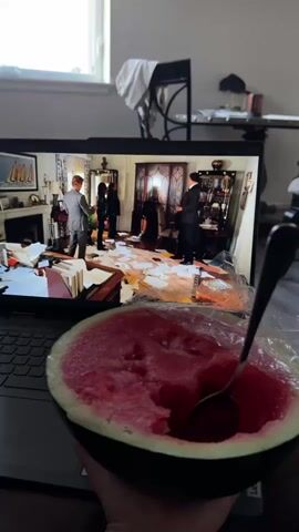 A close-up shot captures a half-eaten watermelon in the foreground, partially covered with plastic wrap, with a spoon resting inside. The watermelon is held in a person's hand. In the background, a laptop screen displays a scene from a movie or show. The screen shows an interior room with people in formal attire, possibly a drawing room or library, with furniture and papers scattered around. The scene then shifts to a close-up of a wooden door with a circular design on it. The overall environment appears to be a dimly lit room, possibly a living room or bedroom, with a window and furniture visible in the far background.