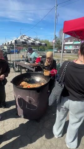 The video shows an outdoor market on a bright, sunny day. Several vendors have set up stalls under canopies. One vendor in the first shot is serving a rice-based dish from a large, black cauldron, while another man grills meat on skewers. Two women stand nearby, presumably waiting for service. The second shot shows the two women speaking to the man at the grill, standing in front of a red canopy. The camera then pans to show a wider view of the market, with other vendors and shoppers moving about. A man on a bicycle moves into the foreground, partially obstructing the view. A red fire extinguisher stands next to the grill. Trees and residential buildings can be seen in the background, adding to the setting. The sky is blue and clear, enhancing the market's lively atmosphere.