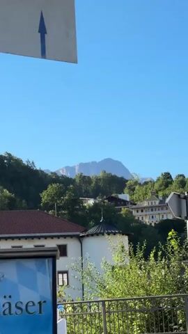 A clear blue sky fills the upper portion of the frame. Below the sky, a mountain range with visible peaks is seen in the distance, partially obscured by a dense, green forest. In the foreground, a white building with a red-tiled roof and several windows is visible. To the right of this building is a smaller, white circular structure with a dark, conical roof. A metal railing runs along the bottom of the frame. A grey signpost with two directional signs is positioned in the middle ground, to the right of center. Lush green foliage and trees are abundant throughout the scene, suggesting a natural, possibly mountainous, environment with some structures integrated into the landscape. The lighting indicates it is daytime.