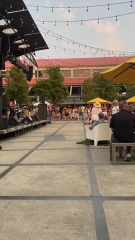 The video shows an outdoor event with a stage set up on the left. A person is performing on stage, possibly singing or speaking into a microphone. There are speakers and sound equipment on the stage. A crowd of people is seated and standing in front of the stage, watching the performance. In the background, there are buildings with awnings and trees. String lights are hung across the open space above the crowd. In the foreground, there are several people sitting on chairs and a bench, facing the stage. One person is sitting in a white chair with a patch of artificial grass underneath. Another person is sitting on a metallic bench. The overall atmosphere appears to be casual and festive, possibly a concert or an outdoor market. The lighting suggests it is late afternoon or early evening.