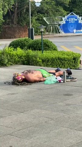 A person lies prone on a paved surface covered in dirt and debris. The person is wearing green shorts and appears to have dirt on their upper body and head. A white and pink bandana is on their head. Next to the person are a blue plastic bag, a silver water bottle, and a pair of shoes. In the background, there are green bushes, a lamppost, and a building with blue signage.