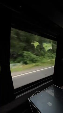 The video shows a view from inside a vehicle, looking out a window at a blurred landscape. The window is framed by black curtains. Outside, a road runs alongside a dense green forest. In the foreground, inside the vehicle, a closed laptop is resting on a surface, with a white cable connected to it. The motion blur outside suggests the vehicle is in motion.