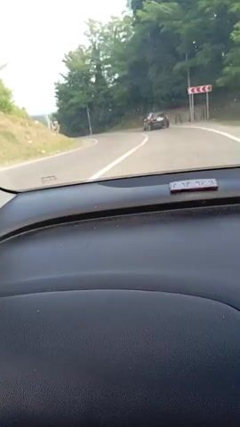 The video shows a view from inside a car, looking out through the windshield at a paved road with a white line dividing the lanes. On the right side of the road, there are trees and a traffic sign indicating a sharp curve to the right. In the distance, there are two black cars driving on the road. The first car is further away and appears to be turning. The second car is closer and is also on the road. The car in which the video is being filmed is driving on the same road. The overall environment appears to be a rural or semi-rural area with a lot of greenery. The sky is visible and appears to be bright, suggesting daylight. The video is shot from a moving vehicle, indicated by the slight blur and the sense of motion.