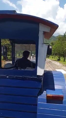 The video shows the interior of a blue and white train engine with a person in a dark shirt and cap operating it. The train is moving along a paved path with trees and green hills in the background. The scenery suggests a park or recreational area. The video is brief and focused on the train's movement and the operator.