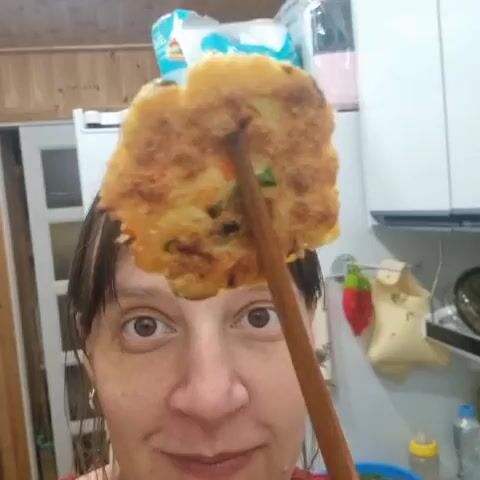 A woman is holding a food item with chopsticks near her mouth. The food item is round and appears to be fried with some green pieces mixed in. The woman is smiling and looking at the camera. The background shows a kitchen with cabinets and a refrigerator.
