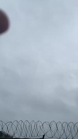 A low-angle shot captures a section of barbed wire fence against a cloudy, overcast sky. The barbed wire is coiled and taut, stretching horizontally across the bottom of the frame. The sky is a uniform gray, with no discernible features or landmarks. The overall impression is one of bleakness and confinement.