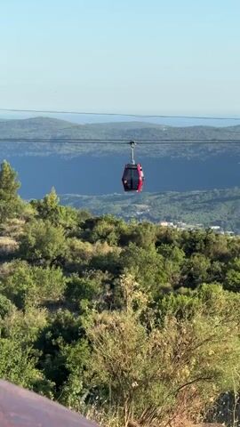 A red cable car moves along a wire suspended over a green, hilly landscape. The sky is a clear blue. In the distance, there are faint outlines of mountains and the sea. The foreground shows a wooden railing.