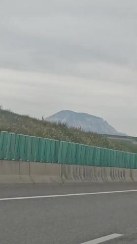 A video captures a view from a moving vehicle on a highway. The camera is pointed forward and slightly to the right. In the distance, a large, flat-topped mountain is visible under a cloudy sky. A green sound barrier runs along the side of the highway, separating it from a grassy embankment dotted with small white flowers. The highway itself is paved, with white lane markings visible. Further down the highway, a few cars and trucks can be seen. The overall environment appears to be rural or semi-rural, with fields and possibly some scattered trees beyond the sound barrier. The lighting suggests it is daytime, but the overcast conditions prevent harsh shadows.