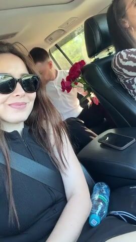 Two women and a man are in a car. The woman in the foreground is wearing sunglasses and a black top, and has a seatbelt fastened. She is smiling at the camera. Behind her, a man is seated in the back seat, holding a bouquet of red roses. He is wearing a white t-shirt and black pants. Another woman is in the driver's seat, wearing a patterned top. A bottle of water and a smartphone are on the center console. The car appears to be in motion, with trees and greenery visible outside the windows.