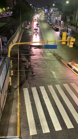 The video shows a street scene at night after a rain. Cars are moving in the distance, and a few pass closer by. There is a crosswalk in the foreground. Traffic lights hang above the street. The street is wet and reflects the light. There is a train track running along one side of the street. Buildings line the other side of the street.