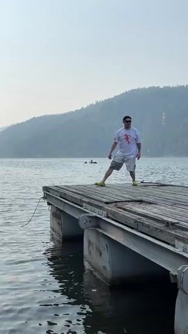 A person stands on a floating dock on a lake. The person is wearing a white t-shirt with a red graphic, grey shorts, and yellow shoes. They are looking towards the camera and have sunglasses on. The dock is made of wood planks and pontoons. In the background, there is a calm body of water, a forested hill, and a hazy sky. There are two small boats or kayaks in the water further out. The lighting suggests it is daytime, possibly late afternoon due to the haze. The person is standing still and the camera is stationary.