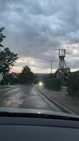This is a first-person view from inside a vehicle driving on a wet road under a cloudy sky. The road appears to be curving to the left. In the distance, two sets of headlights are visible, indicating oncoming traffic. On the right side of the road, there is a tall, metallic tower structure, possibly an old mining headframe, with a fenced area around its base. A dense hedge runs along the side of the road next to the tower. On the left side of the road, there are trees, a streetlight, and a house. The overall atmosphere is overcast and slightly gloomy, suggesting it might be evening or that rain has recently fallen. There are no visible people in the scene.