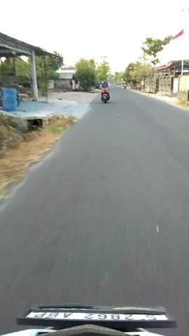 The video shows a first-person perspective of someone riding a motorcycle down a paved road. In the distance, several other motorcycles are also visible, as are trees, buildings, and what appear to be small shops or residences lining the street. One of the flags visible appears to be red and white. The road is in good condition, and the weather seems clear and sunny. The overall impression is of a typical, everyday scene in a populated area.