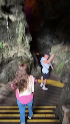 A group of people walks through a narrow cave with wooden steps and walkways. The cave walls are rugged and vary in color from gray to brown, with some green moss growing on them. Overhead, the cave ceiling is lit with orange and purple lights. The people walk single file, with a woman in a pink shirt and blue jeans leading the way. A man wearing a white tank top, black shorts, and a baseball cap follows close behind. Two other people are visible further into the cave, one in a white shirt and one in a tan shirt.