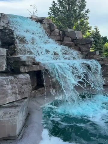 A bright blue, artificial waterfall cascades down a multi-tiered rock structure in Chelsea, Canada. The water, dyed a vibrant aqua color, flows vigorously over large grey boulders, creating foamy white splashes at each level. Green foliage, including tall trees and smaller shrubs, frames the left side of the waterfall. The sky above is a clear blue with scattered white clouds, indicating a daytime scene. The overall impression is one of a constructed natural-looking feature in a park or garden setting.
