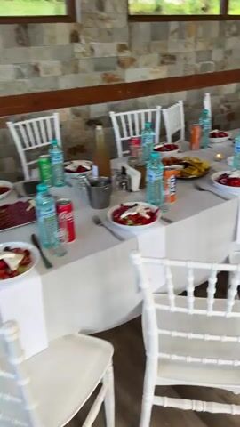A long, white-clothed table is set for an event with white chairs on both sides. Green bottles of water and red and orange sodas are on the table, along with small plates of what appears to be salad with white cheese and red onions, and larger plates of various snacks like cheese and olives. A tall glass vase with green leaves is positioned at the far end of the table. The wall behind the table is made of stone.
