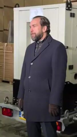 The video shows two men standing in a large, open warehouse space. Stacks of packed wooden planks are visible in the background.  The man on the left has brown hair and a beard and is wearing a dark overcoat, vest, striped pants, and dark brown gloves. He appears to be standing next to a generator sitting on a trailer. The man on the right is younger, and wears a dark hooded jacket, black jeans, and dark shoes. The trailer features red tail lights and a white license plate. Both men are looking off-screen as one speaks.