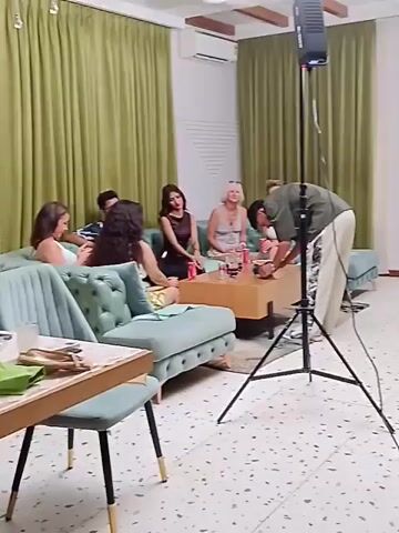 The video shows a scene indoors, likely a living room, where a group of people are gathered. Several women are seated on a teal-colored sectional sofa around a coffee table, while two men are present, apparently setting up lighting equipment. One man is adjusting items on the coffee table, while the other is standing near a door, looking at a smartphone. There's a large black light stand in the foreground. The room is decorated with green curtains and the floor has a speckled pattern.  The ceiling has a fan and exposed wooden beams.