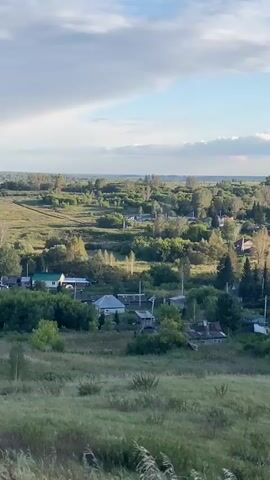 The video displays a panoramic view of a rural landscape under a cloudy sky. The scene transitions from a foreground of tall, dry grass and weeds to a mid-ground and background consisting of rolling hills covered in lush green trees and vegetation. Scattered throughout the landscape are small, dispersed clusters of houses, identifiable by their roofs and structures. Power lines are visible in several areas, connecting the scattered dwellings. The overall impression is that of a tranquil, undeveloped countryside dotted with small villages or hamlets. The lighting suggests it is either early morning or late afternoon, with the sun casting a warm glow on the scene. No people or animals are discernible.