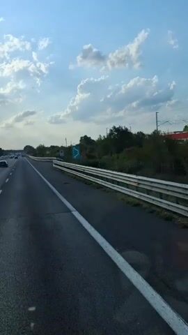 A first-person perspective video of traveling on a highway. The sky is partly cloudy with the sun shining through. There is traffic on the highway, including cars and trucks. The video captures the road ahead, with a guardrail on the right side and a white line marking the lane. There are some green signs on the right side of the road. The video appears to be shot from inside a vehicle.