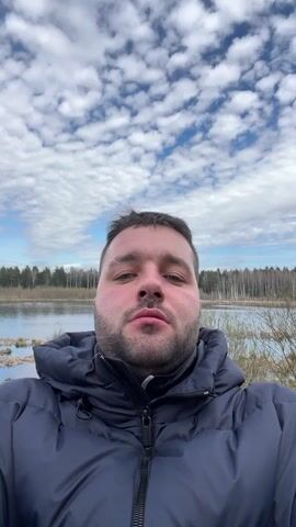 The video shows a man taking a selfie outdoors. He is wearing a dark blue puffer jacket with the hood up. He has short brown hair and a short beard. The background includes a body of water, likely a lake or river, with trees visible on the opposite shore. The sky is bright with many fluffy white clouds. The man looks directly at the camera with a slight smile, his expression generally calm. In the background, there is faint children's speech audible.