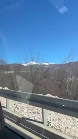 The video shows the view from inside a vehicle moving along a paved road, with a clear blue sky above. A gray metal guardrail is visible on the left side of the road. Beyond the guardrail, the terrain is rocky and sparsely vegetated. In the distance, there are dark brown mountains topped with snow. The windshield has some reflections on it. The video is shot in bright daylight. There is music playing.