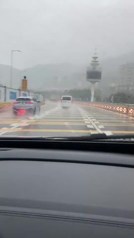 A car is driving on a wet road during a downpour. The windshield wipers are active, clearing raindrops. Other vehicles, including a silver car and a white van, are visible ahead on the road. A tall, distinctive tower with a circular observation deck stands to the right of the road. Mountains and buildings are visible in the background, obscured by rain and fog. The road has multiple lanes marked with yellow and white lines. The sky is overcast, indicating heavy rain. The perspective is from inside the driving car.