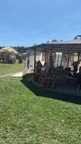 The video shows an outdoor market scene on a sunny day. In the foreground, a well-maintained green lawn is visible. In the middle ground, a covered stall is set up with a corrugated metal roof and open sides, displaying various items, possibly for sale. There are several people in and around the stall. To the left of the stall, there is a white tarp extending outwards. In the background, other market stalls with awnings and more people can be seen, with a line of trees in the distance against a clear blue sky. The overall atmosphere suggests a lively outdoor event.