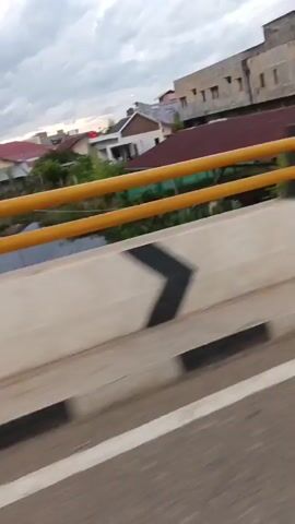 The video is taken from the perspective of someone riding on the back of a motorcycle. The motorcycle is moving along a road, which appears to be in a town or city. There are buildings on either side of the road, and several other motorcycles are also on the road. In the foreground, the hands of the rider are visible, as is the back of the head of the person driving the motorcycle. The rider in the back is wearing a red shirt. The sky is cloudy and appears to be overcast. Several Indonesian flags are visible, suggesting a national holiday or event. The road has a white line marking the lane and a concrete barrier with black and yellow chevron markings. Traffic lights are visible in the distance, and some appear to be red. The overall scene depicts movement and activity within a populated area.