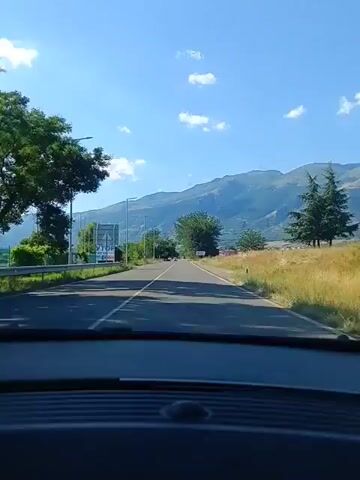The video shows a first-person view from inside a car driving down a paved road. The sky is blue with scattered white clouds. In the distance, there are mountains covered in green trees. On the sides of the road, there are grassy fields, trees, and streetlights. There are a few other cars visible on the road ahead, and a billboard on the left side. The dashboard of the car is visible in the foreground. The overall impression is of a scenic drive on a sunny day.
